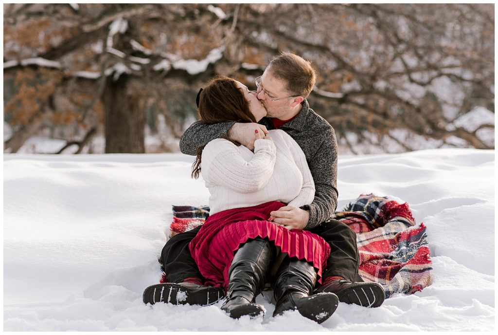 winter engagement photos, red and white engagement outfit inspiration, sitting in snow photos, cozy engagement photos, romantic engagement photos, plaid blanket engagement prop, plaid blanket prop, engagement session props