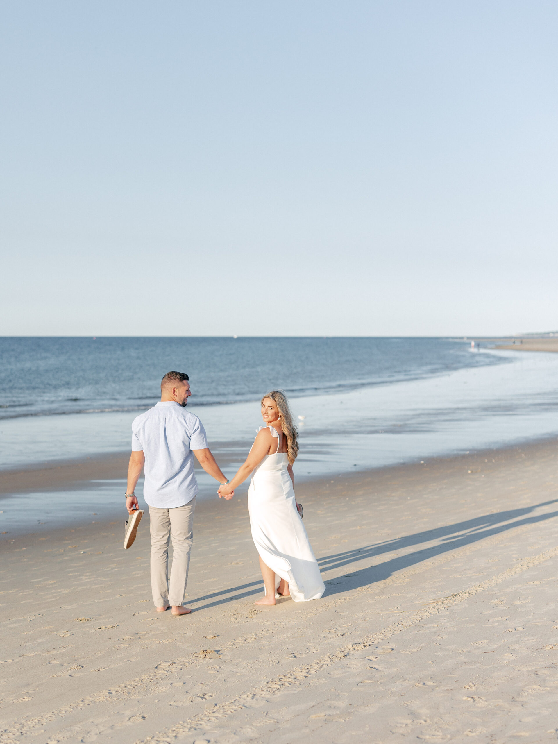 Guy and girl hold hands while walking with shoes in hand down the beach and the girl is looking back at the camera while the guy looks at the girl