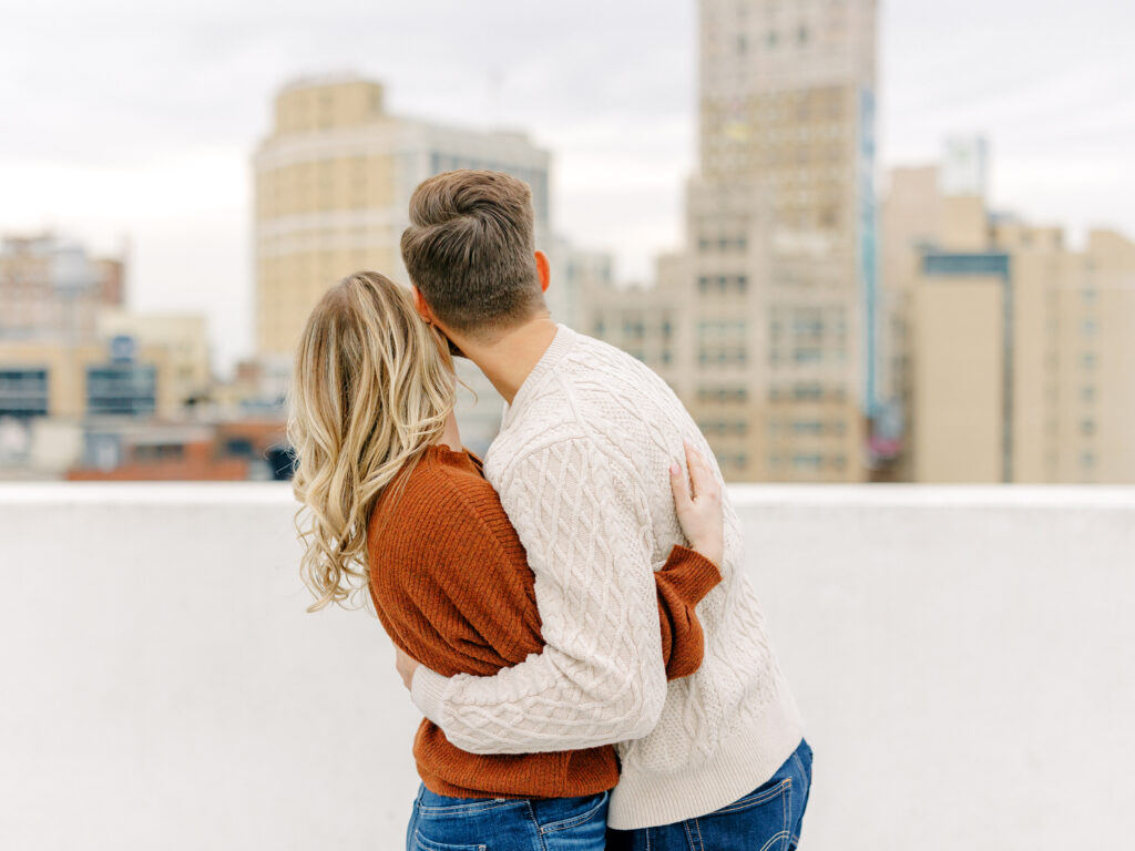 Caroline and Nick embracing on a rooftop parking garage with the Detroit skyline in the background.